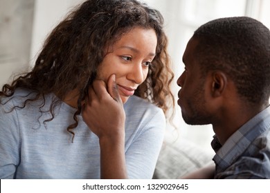 Loving African American man cheering beloved woman touching her face relaxing together at home, caring black boyfriend support mixed race girlfriend making her smile, looking in eyes