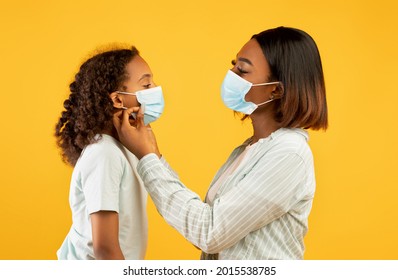 Loving African American Woman Putting Medical Mask On His Cute Daughter Face, Yellow Background. Mother And Girl Going Out During COVID-19 Pandemic, Parent Helping Child To Wear Protective Face Mask