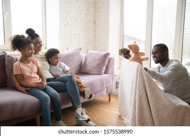 Loving african American young dad make in puppet show to excited young wife sit relax on couch with little kids, happy biracial family have fun entertain in cozy living room, watch theater play