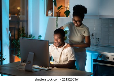 Loving african woman massaging shoulders, doing cervical massage for husband while he working late on computer at home, black guy developer writing code on computer at night with wife next to him