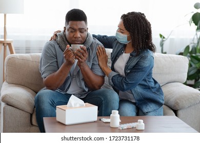 Loving Afro Woman In Medical Mask Taking Care About Her Sick Husband Suffering From Coronavirus At Home, Offering Him Tea, Free Space