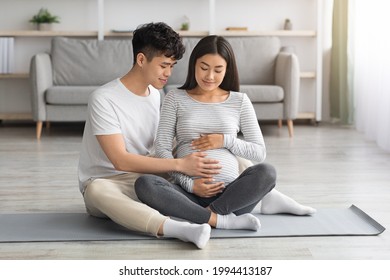 Loving Asian Man Husband Touching His Pregnant Wife Big Belly, Copy Space. Cheerful Chinese Expecting Family Enjoying Healthy Lifestyle During Pregnancy, Doing Sport At Home, Cuddling, Embracing