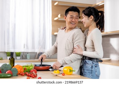 Loving Asian Middle Aged Man Husband Preparing Delicious Healthy Meal For His Wife, Happy Young Chinese Lady Hugging Her Spouse Cooking Dinner, Kitchen Interior, Side View, Copy Space