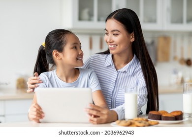 Loving Asian Mother And Her Daughter Using Digital Tablet, Looking For New Recipes While Sitting At Kitchen Table And Enjoying Cookies With Milk. Young Mom And Child Spending Time Together