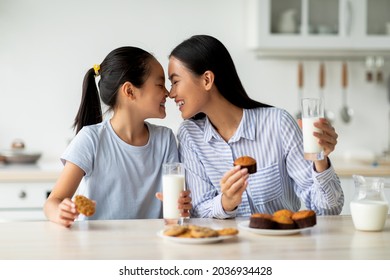 Loving Asian Mother And Little Daughter Having Snacks And Drinking Milk In Kitchen Interior, Enjoying Freshly Baked Cakes. Happy Family Eating Homemade Pastry And Bonding At Home