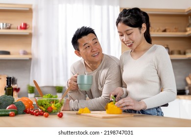 Loving Asian Young Woman Wife Preparing Delicious Healthy Meal For Her Husband, Happy Middle Aged Chinese Man Drinking Tea Next To His Spouse Cooking Dinner, Kitchen Interior, Copy Space