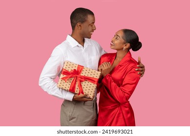 Loving black couple exchanging gifts, with man giving wrapped present to smiling woman in red dress, expressing affection and happiness - Powered by Shutterstock