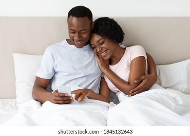 Loving Black Couple Holding Mobile Phone, Watching Photos Or Videos Together, Lying On Bed At Home, Copy Space. African American Young Man And Woman Using Smartphone In Bedroom, Having Fun Together