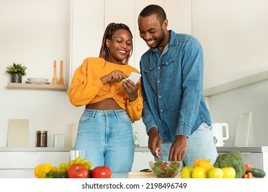 Loving Black Couple Reading Food Blog On Smartphone, Cooking Together Healthy Vegetable Salad At Home Kitchen. Happy Man And Woman Making Lunch, Checking Recipes On Internet