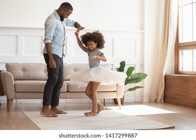 Loving black dad holding hand of child daughter dancing with his child princess wearing skirt, happy young african father and little cute daughter playing in living room enjoy time together at home - Powered by Shutterstock