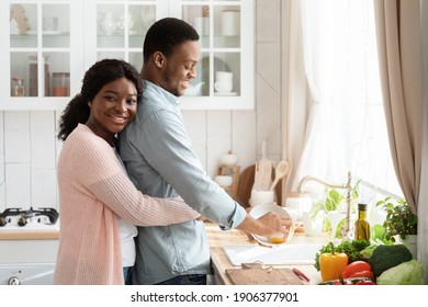 Loving Black Husband Helping With Cleaning, Washing Dishes After Lunch In Kitchen. Affectionate African Wife Hugging Her Spouse From The Back And Smiling, Grateful For Assistance In Domestic Chores