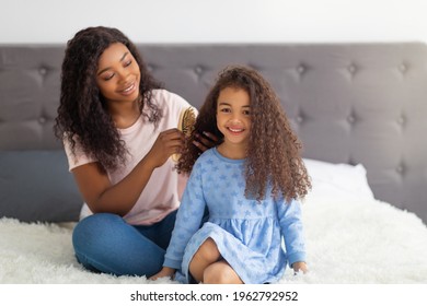 Loving black mom brushing her pretty daughter's hair on bed at home. Cute little child and caring mother doing hairstyles, having fun together, playing beauty salon, spending spa day indoors - Powered by Shutterstock