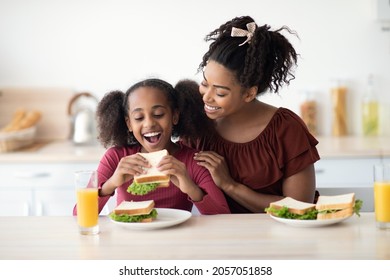 Loving Black Mother And Daughter Cuddling While Having Snack At Kitchen, Eating Healthy Sandwiches And Drinking Orange Juice, Happy Mom Embracing Her Hungry Kid Teen Girl, Copy Space