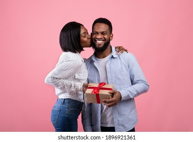 Loving Black Woman Giving Her Happy Husband Valentine Gift, Kissing Him On Cheek Over Pink Studio Background. Emotional African American Couple Celebrating Lovers Holiday Together, Exchanging Presents