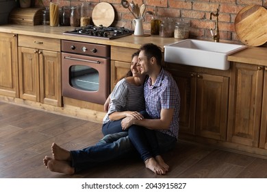 Loving Bonding Young Latin Family Couple Sitting On Warm Heated Wooden Floor In Kitchen, Daydreaming Looking In Distance, Visualizing Future Or Enjoying Sweet Leisure Weekend Moment In Own House.