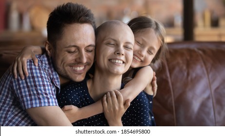 Loving and caring. Affectionate happy young husband dad and school age daughter kid sitting on couch supporting cuddling warm tight beloved smiling millennial wife mom fighting against cancer disease