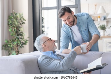 Loving caring young adult son bringing his old elderly senior father a cup of water hot drink at home indoors. Elderly generation support and help aid. Happy father`s day! I love you, dad!