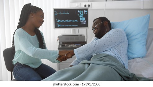 Loving And Caring Young African Woman Visiting And Talking To Hospitalized Husband. Afro-american Male Patient Lying In Hospital Bed, Holding Hands And Chatting To Wife Visitor