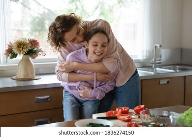 Loving Caucasian Mom Hug Play With Teen Daughter Cooking Tasty Salad For Dinner At Home Kitchen. Caring Happy Mother Enjoy Day With Teenage Girl Child, Laugh Have Fun Preparing Healthy Food Together.