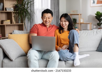 Loving Cheerful Chinese Couple Using Laptop Together, Cozy Home Interior, Happy Asian Man Hugging His Beautiful Wife And Looking At Notebook Screen, Watching Photos, Copy Space