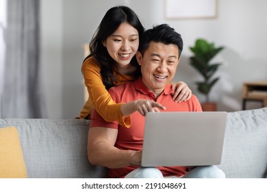 Loving Chinese Spouses Shopping On Internet From Home, Cheerful Young Asian Woman Hugging Her Husband From Behind, Pointing At Laptop Screen, Family Choosing New Furniture Online, Copy Space