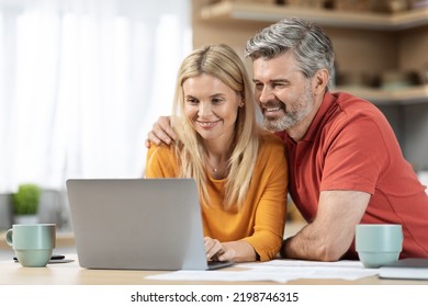 Loving Couple Attractive Middle Eastern Man And Woman Sitting At Kitchen Table In Front Of Computer, Looking At Laptop Screen, Hugging And Smiling, Paying Bills On Internet, Copy Space