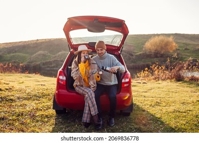 Loving couple chilling in car trunk by autumn lake drinking tea from vacuum flask. Man and woman travel by auto. Young people enjoy fall activities