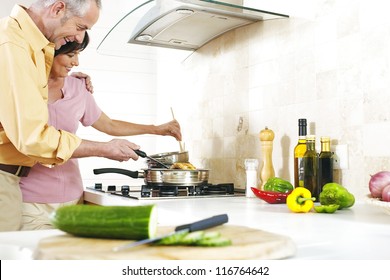 Loving Couple In A Close Embrace Stand Over The Gas Hob Preparing A Meal In A Metal Frying Pan