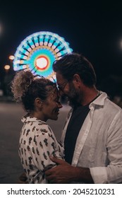 Loving Couple Embracing Each Other Passionately On Street Against Illuminated Ferris Wheel At Night. Couple Romancing And Spending Quality Time Outdoors During Vacation At Night