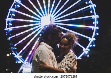 Loving Couple Embracing Each Other Passionately On Street Against Illuminated Ferris Wheel At Night. Couple Romancing And Spending Quality Time Outdoors During Vacation At Night