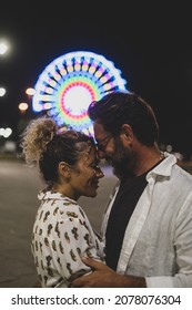 Loving Couple Embracing Each Other Passionately On Street Against Illuminated Ferris Wheel At Night. Couple Romancing And Spending Quality Time Outdoors During Vacation At Night