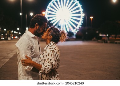 Loving Couple Embracing And Kissing Each Other Passionately On Street Against Illuminated Ferris Wheel At Night. Couple Romancing And Spending Quality Time Outdoors During Vacation At Night