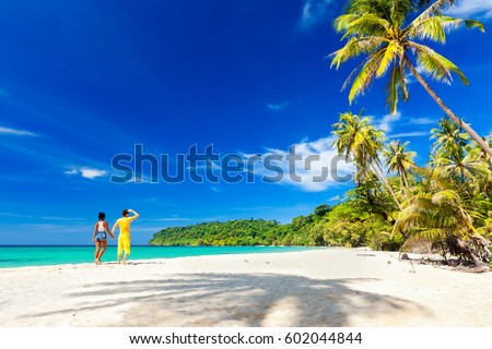 A loving couple enjoying the breathtaking views of the tropical sandy beach with green coconut palm trees and beautiful clean sea ocean on a background of blue sky