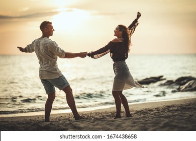 A loving couple is having fun and dancing on the empty sandy sea beach at the sunset.They are looking each other and happily smiling.