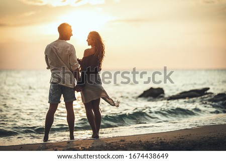 A loving couple is having fun and hugging on the empty sandy sea beach at sunset.They are looking each other and happily smiling.