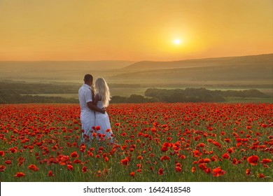 Loving Couple Hug One Another During Romantic Date In Marvellous Spring Poppy Field With Bright Sunset Above Forest And Mountains. People In Wild Nature Landscape Horizontal Background.