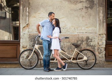 Loving Couple Hugging Near The Tandem Bike Against Old Wall On Street In Town. Handsome Man In Blue Shirt Is Kissing In Head Woman Wearing White Dress