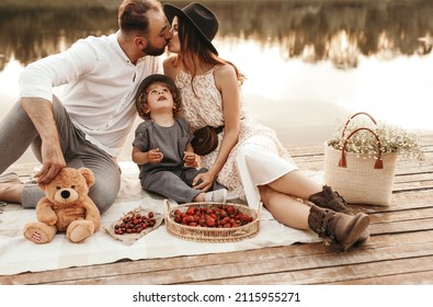 Loving couple kissing and sitting on blanket with little child while having picnic with fresh berries on wooden quay near pond in summer