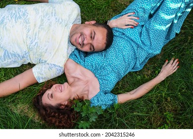 Loving couple lie on the grass look at each other and laugh. Man in a T-shirt and a curly woman in a blue dress on a romantic date. A Middle Eastern man with his caucasian wife.