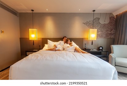 Loving Couple Of Man And Woman Lying And Relaxing Together On Bed In Luxury Hotel. Romantic Moments Of Two Lovers On Holidays Or Vacation In Modern Resort. Family Honeymoon Trip Or Travel