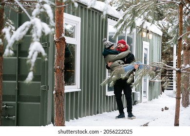 Loving Couple Outdoors At Winter. Man Is Carrying His Woman In His Hands. Both Wearing Hats, Jackets And Scarfs. In Front Of A House With Corrugated Sheet Wall.