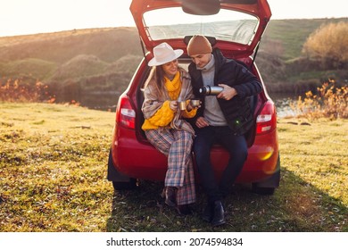 Loving couple relaxing in car trunk by autumn lake drinking tea from vacuum flask. Man and woman travel by auto. Young people enjoy fall activities