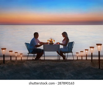 Loving couple share a romantic dinner with candles, lanterns and wine glasses at sea beach sand against wonderful sunset