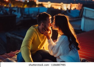 Loving couple is sitting on the roof of the house. In the distance, the lights of the night city.