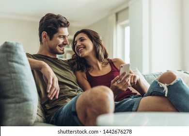 Loving couple sitting on sofa at home. Man and woman relaxing on couch with smartphone in living room.