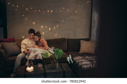 Loving Couple Sitting On Sofa And Using Mobile Phone At Home. Man And Woman Relaxing On Couch With Smartphone In Cozy Living Room.