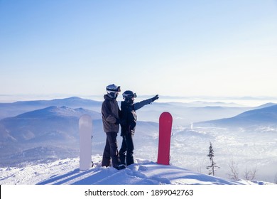 Loving couple of snowboarders hold hands and look forward, man indicates direction to woman with snowboard route with hand.