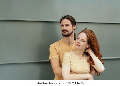 Loving Couple Standing Watching To The Side With Serious Intent Expressions In A Close Embrace Against A Grey Exterior Wall