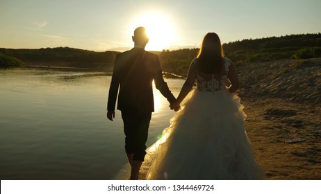 loving couple walks hand in hand on the summer beach in the rays of sunset. The bride in white dress and the groom barefoot walking on the water on the river bank. Slow motion.