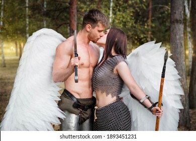 A loving couple of warrior angels kissing. Athletic man and woman in armor with weapons in their hands and white wings behind their backs against the backdrop of the park.